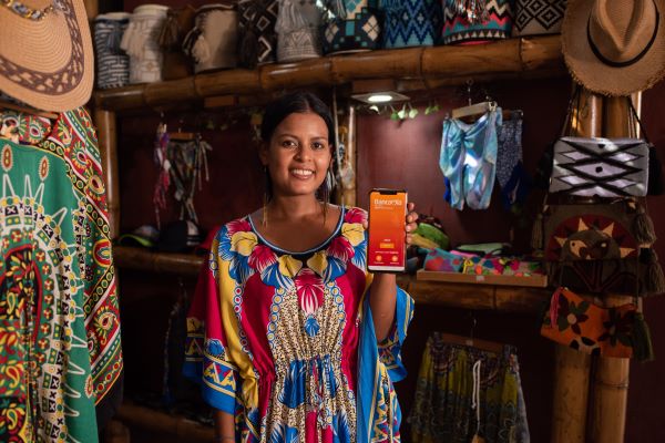 Mujer con un vestido de colores sonriendo en una tienda de artesanias, mostrando la app Bancamia desde el celular
